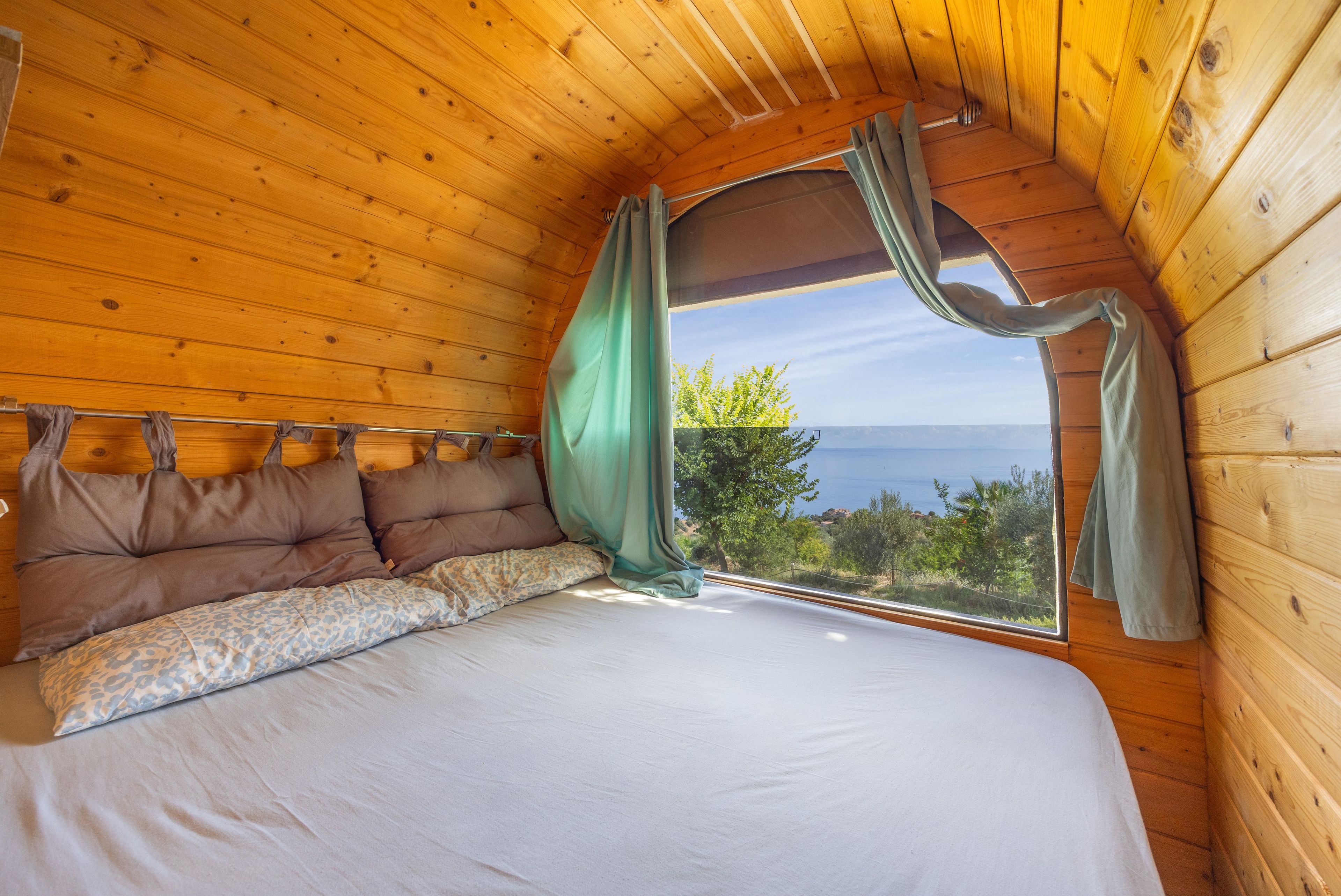 Un dormitorio acogedor con techo de madera, una cama con colchón blanco y almohadas estampadas, y una ventana con cortinas.