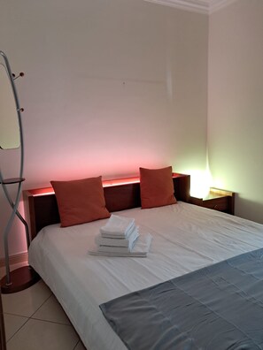 Ein ordentlich bezogenes Bett mit weißen Leinen und einem gestapelten Handtuchset, zwei rote Kissen, ein Nachttisch mit einer Lampe und ein metallischer Klappstuhl.