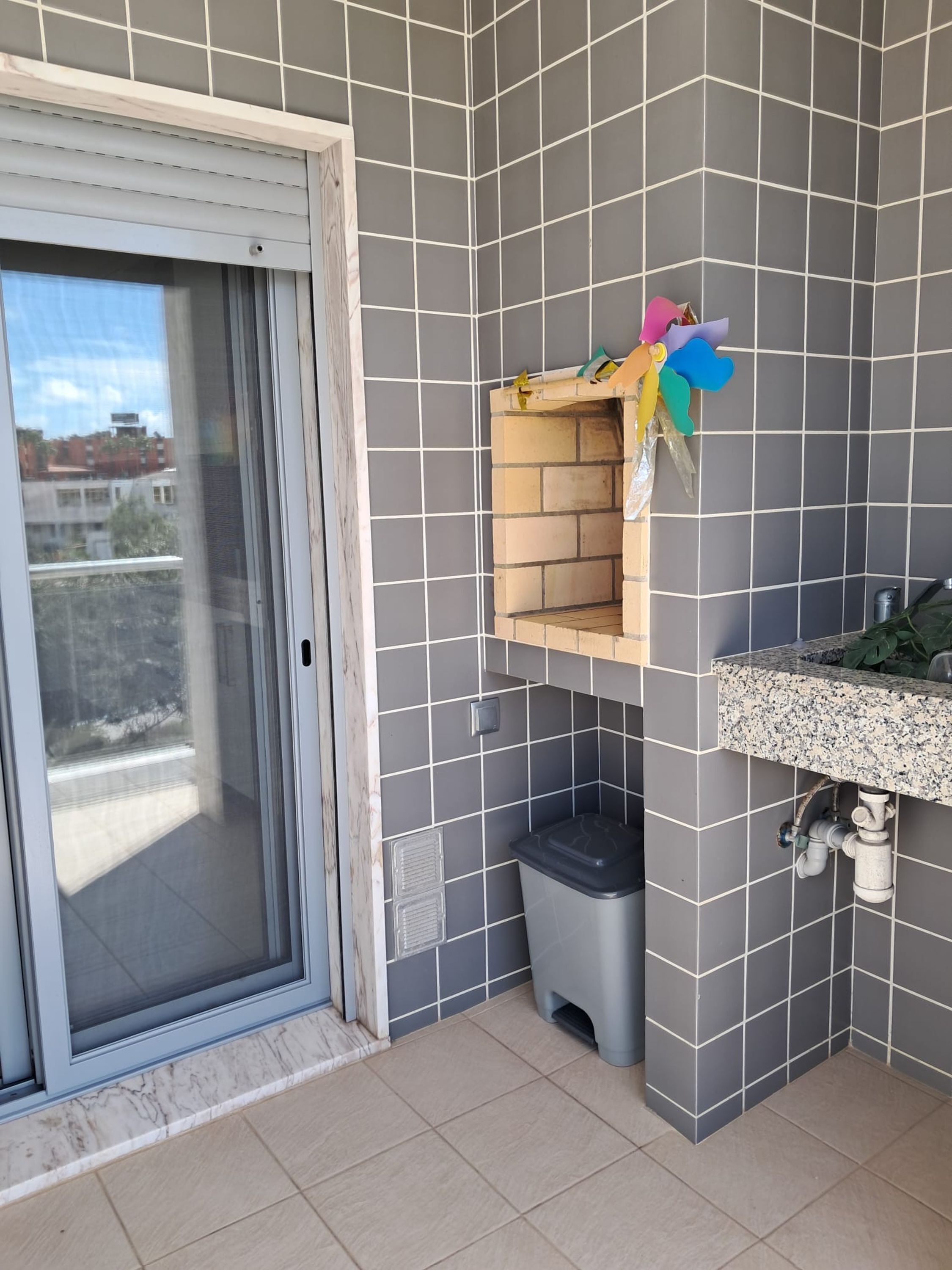 Un balcon avec des murs recouverts de carreaux gris, une porte-fenêtre coulissante, une petite poubelle et une étagère ornée de décorations colorées.