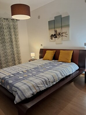 Ein Schlafzimmer mit Bett, Nachttisch, Lampe, einem Fenster mit Vorhängen und Wandbildern.