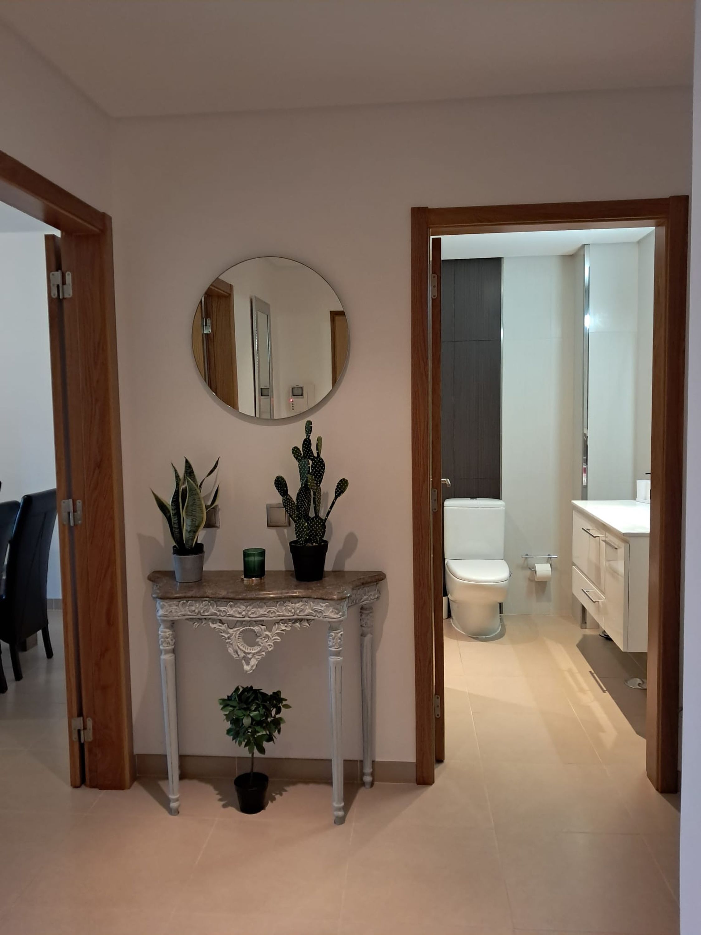 Ein Flur mit einem runden Spiegel, einem Konsolentisch mit Pflanzen und Blick ins Bad mit Toilette und Waschbecken.