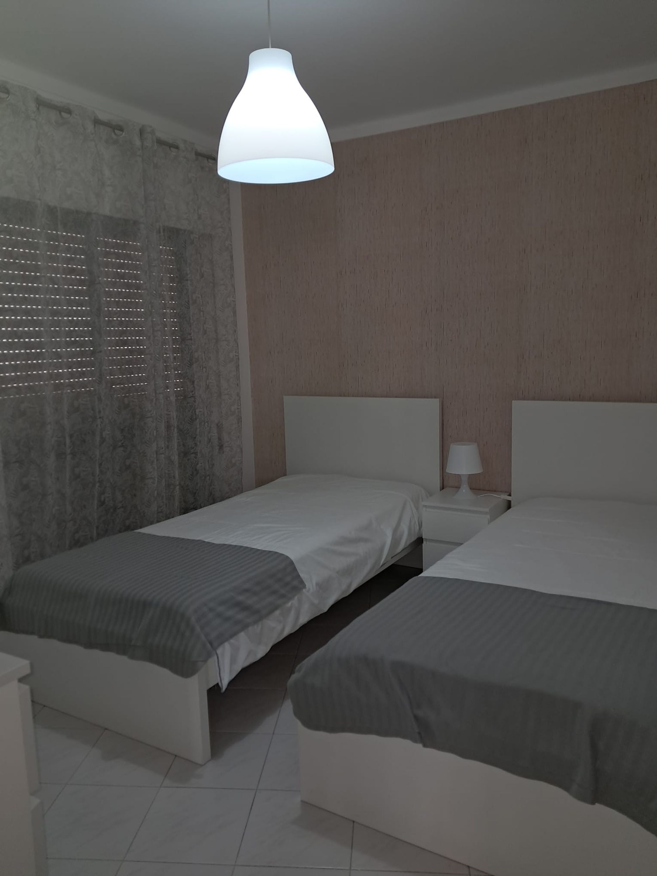 Ein Hotelzimmer mit zwei Einzelbetten, einem Nachttisch mit Lampe, einem Fenster mit Jalousien und einer Pendelleuchte.