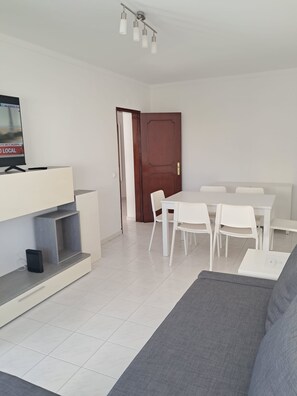 A modern living room with a sofa, a dining table with chairs, a TV on a stand, and a wooden door. - T2 Vista MAR in Quarteira (Faro)