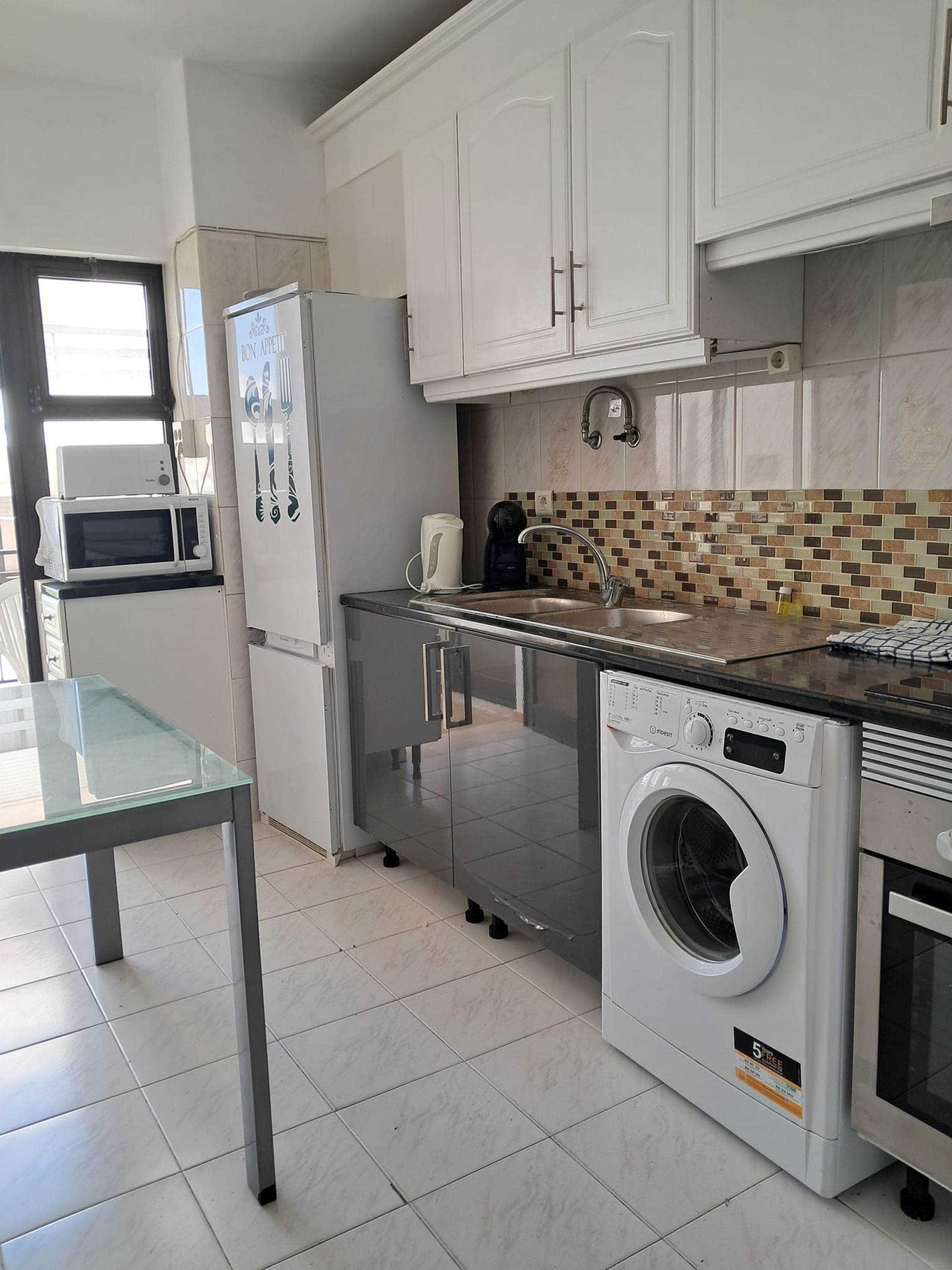 A compact kitchen with a microwave, refrigerator, coffee maker, washing machine, and a glass table.