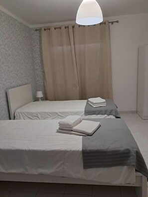 A hotel room with two beds, a headboard, a bedside table with a lamp, and a large window with curtains. - T2 Vista MAR in Quarteira (Faro)