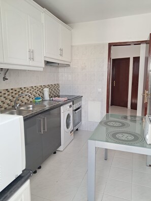 A kitchen with white cabinets, a washing machine, a microwave, and a glass-top table. - T2 Vista MAR in Quarteira (Faro)