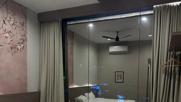 Quarto de hotel moderno com cama, escrivaninha, cadeira, divisória de vidro e ventilador de teto.