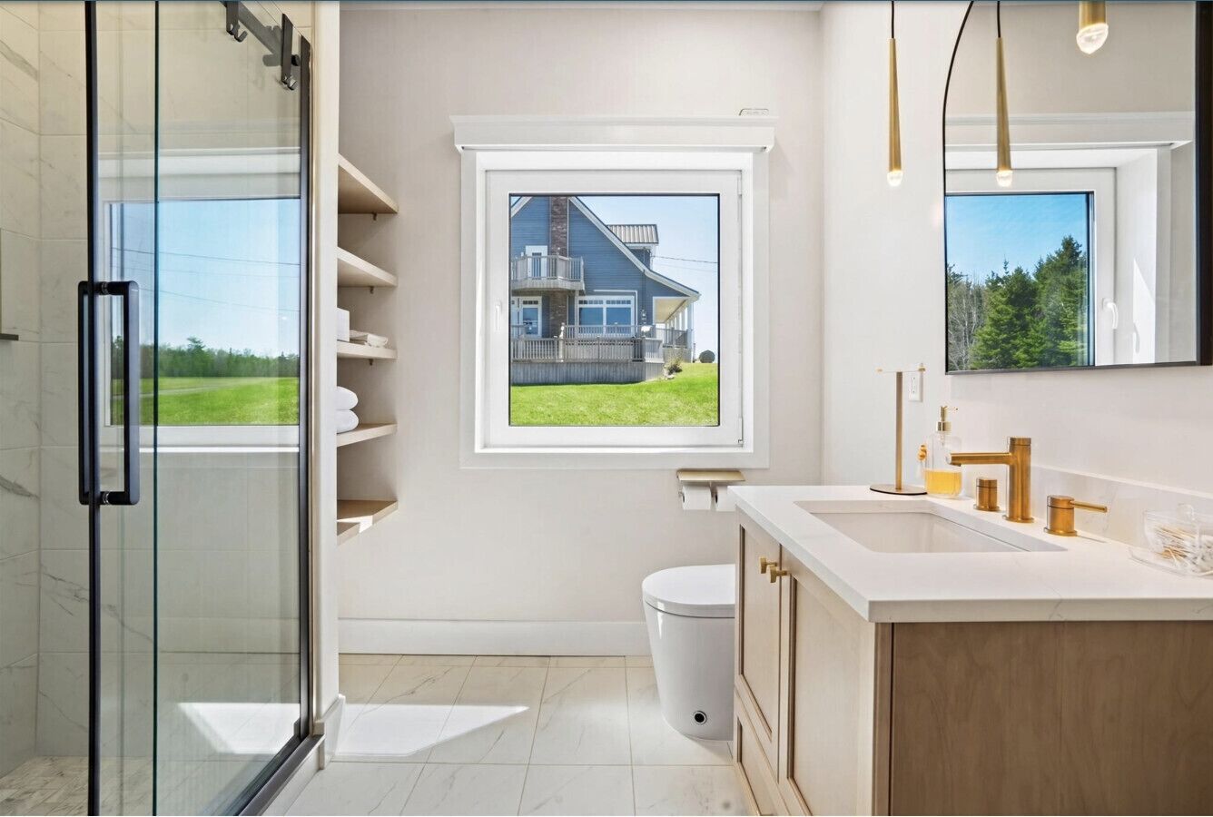 Ein modernes Badezimmer mit Duschkabine aus Glas, Toilette, Waschbecken mit goldfarbenen Armaturen und einem Fenster mit Blick auf ein Haus und Grünflächen.