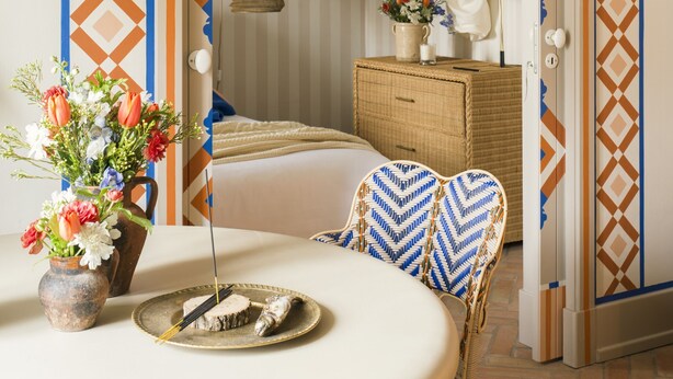 A room with a bed, a wicker chair, a table with a vase of flowers, and a decorative wall pattern. - Al Porta Townhouse by Cadenzalux (Beja)