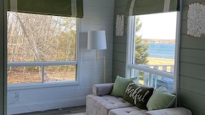 A cozy room with a grey sofa, a green throw pillow, a white lamp, and a view of the outdoors. - Serene Cove Retreat Green Cottage-overseeing beautiful Montague River and and peaceful harbor (Montague)