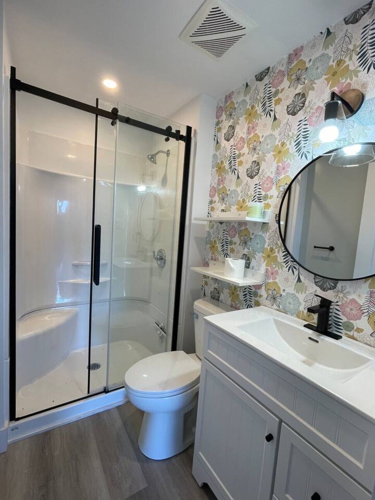 Un bagno con doccia vetrata, un water, un lavandino con specchio e una parete decorata con motivi floreali.
