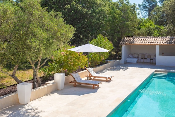 Un espace aménagé au bord de la piscine, avec des chaises longues, un coin salon abrité et une piscine.