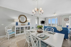 A dining area with a white table, white chairs, a chandelier, and a blue couch in the background.