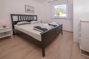 Ein Einzelbett mit Kopfteil, ein Nachttisch mit Lampe, ein Fenster mit Vorhängen und ein weißer Kleiderschrank.