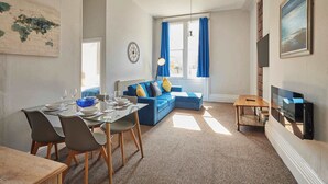En stue med en blå sofa, et spisebord med stole, et ur og et fjernsyn.