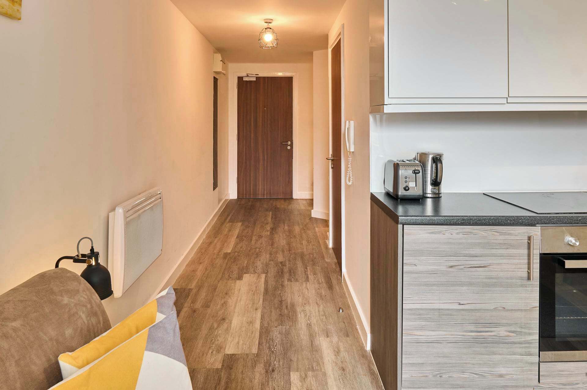 A hallway with a wooden floor, a door at the end, and a kitchenette with a toaster and kettle.