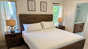 Un dormitorio con una cama de madera, dos mesitas de noche con lámparas, un tocador, un ventanal con cortinas y un baño visible a través de una puerta abierta.
