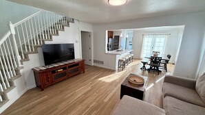 Una sala de estar con un mueble de madera para centro de entretenimiento, un televisor, una escalera, un área de comedor con una mesa y sillas, y una cocina con un fregadero y taburetes.