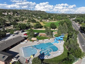 An aerial view of a swimming pool complex with multiple water slides, surrounded by trees and a parking area.