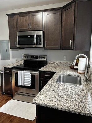 Una cocina con gabinetes de madera oscura, un microondas de acero inoxidable, horno empotrado, encimera de granito y un lavaplatos de acero inoxidable.