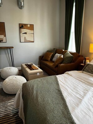 Una habitación de hotel con una cama, un sofá de cuero marrón, dos otomanas, una mesita con una bandeja y obras de arte en las paredes.
