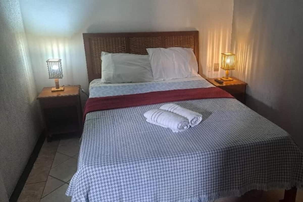 Una cama bien hecha con una colcha a cuadros, una toalla enrollada y dos lámparas de noche.