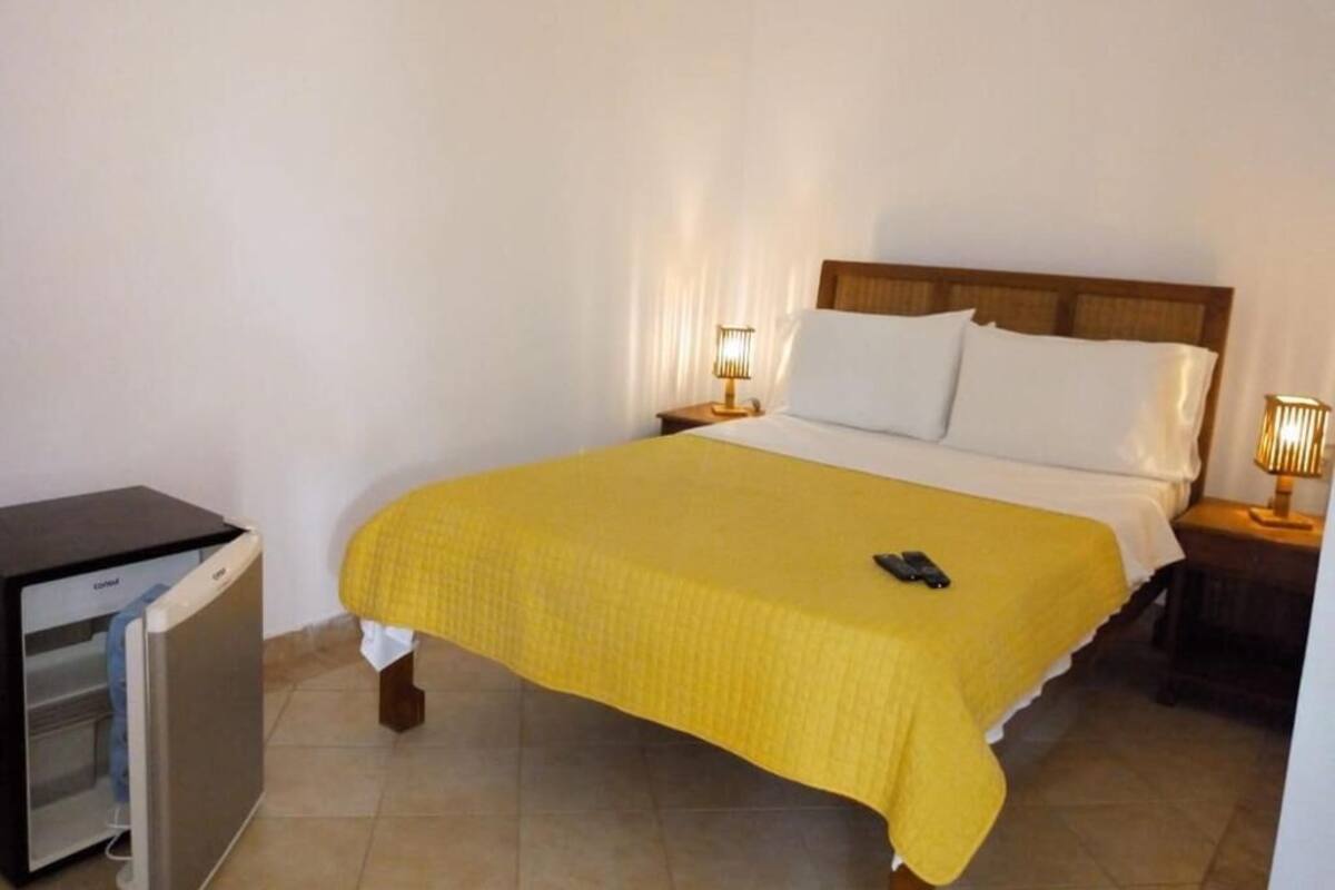 Una cama individual con colcha amarilla, una lámpara de noche y un refrigerador pequeño.