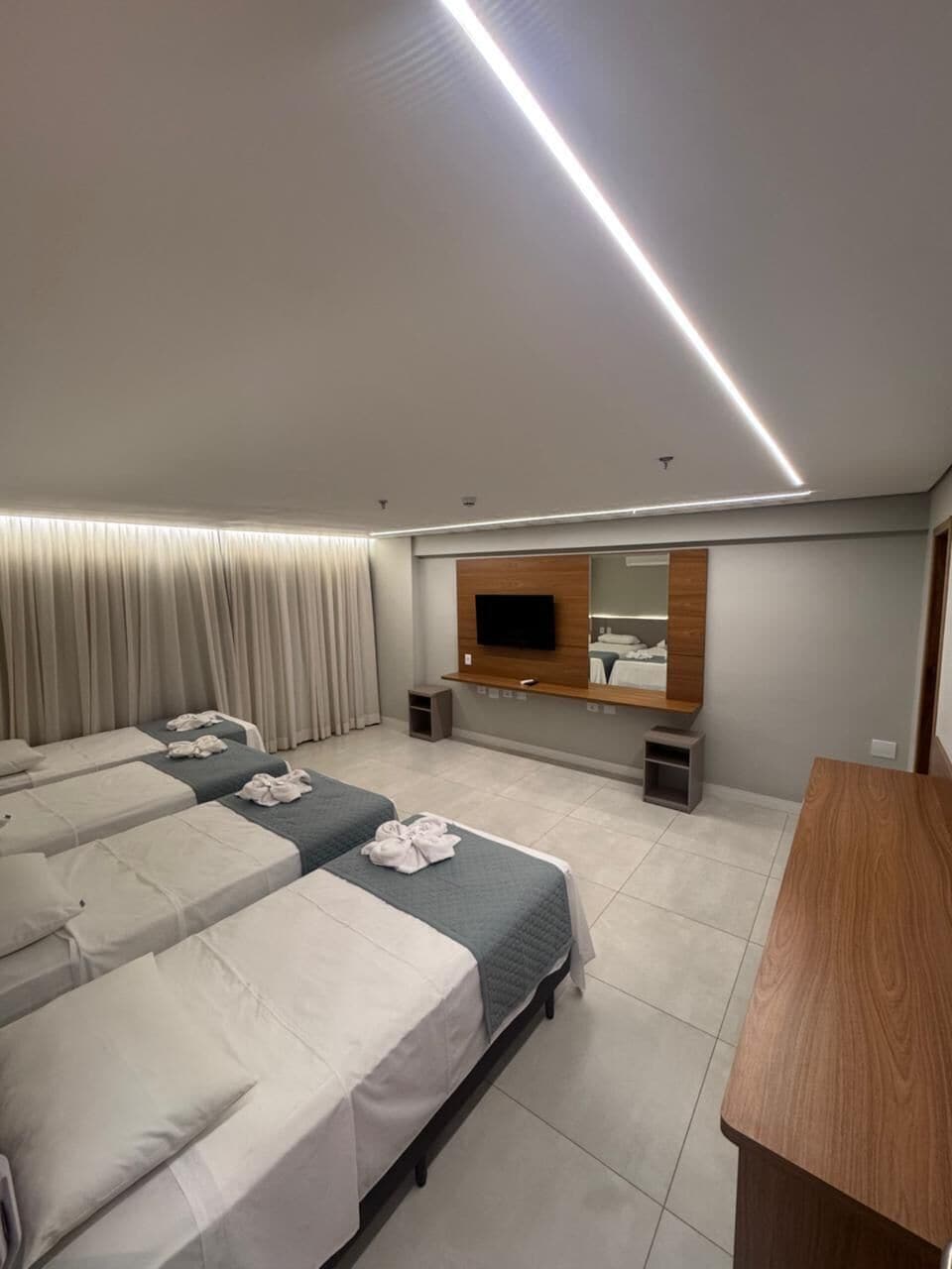 Hotellihuone, jossa on kolme sänkyä, televisio, peili ja puinen työpöytä.