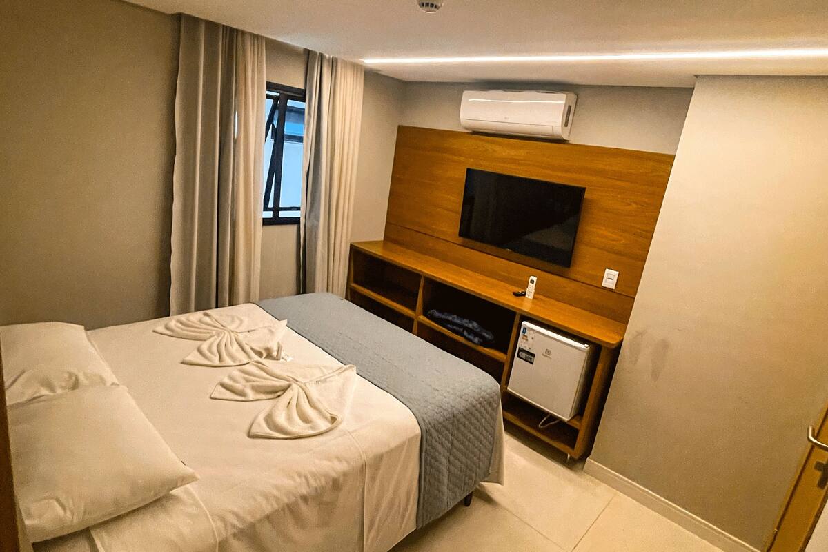 Habitación de hotel con cama, televisor, refrigerador pequeño y ventana con cortinas.