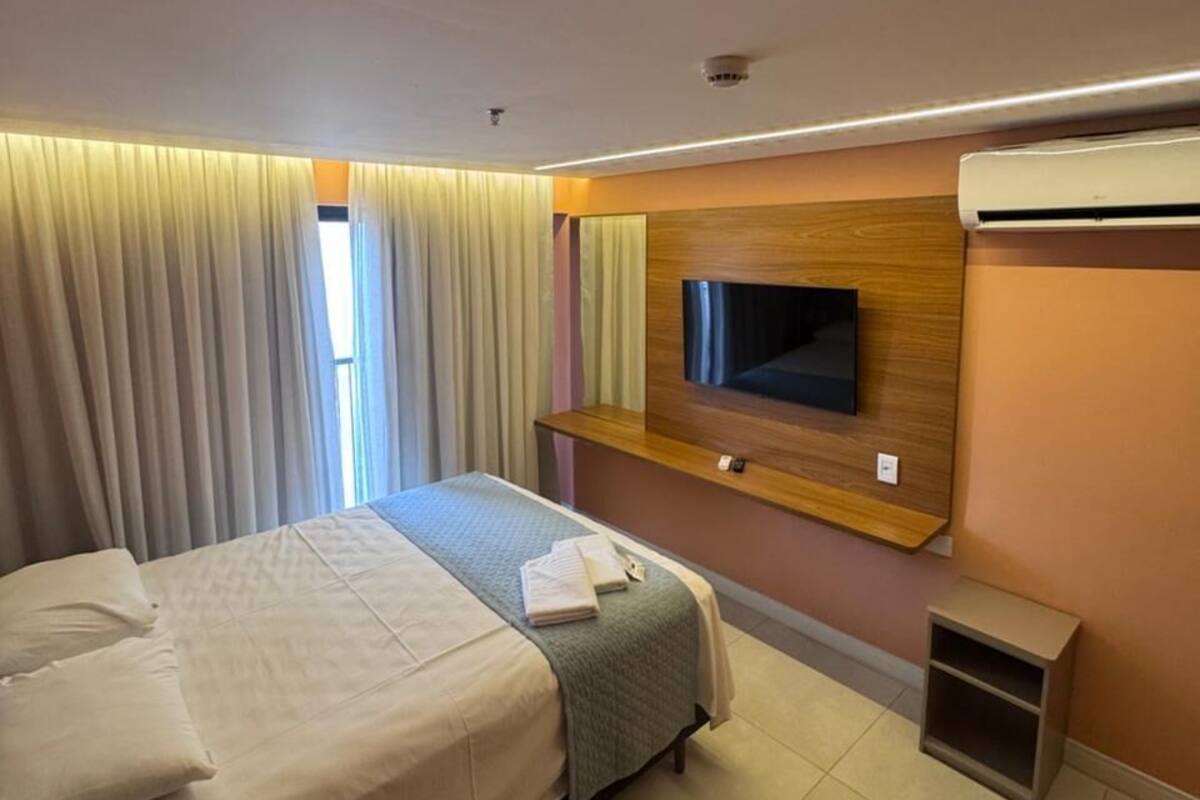 Habitación de hotel con cama, televisión montada en la pared, equipo de aire acondicionado y ventana con cortinas.