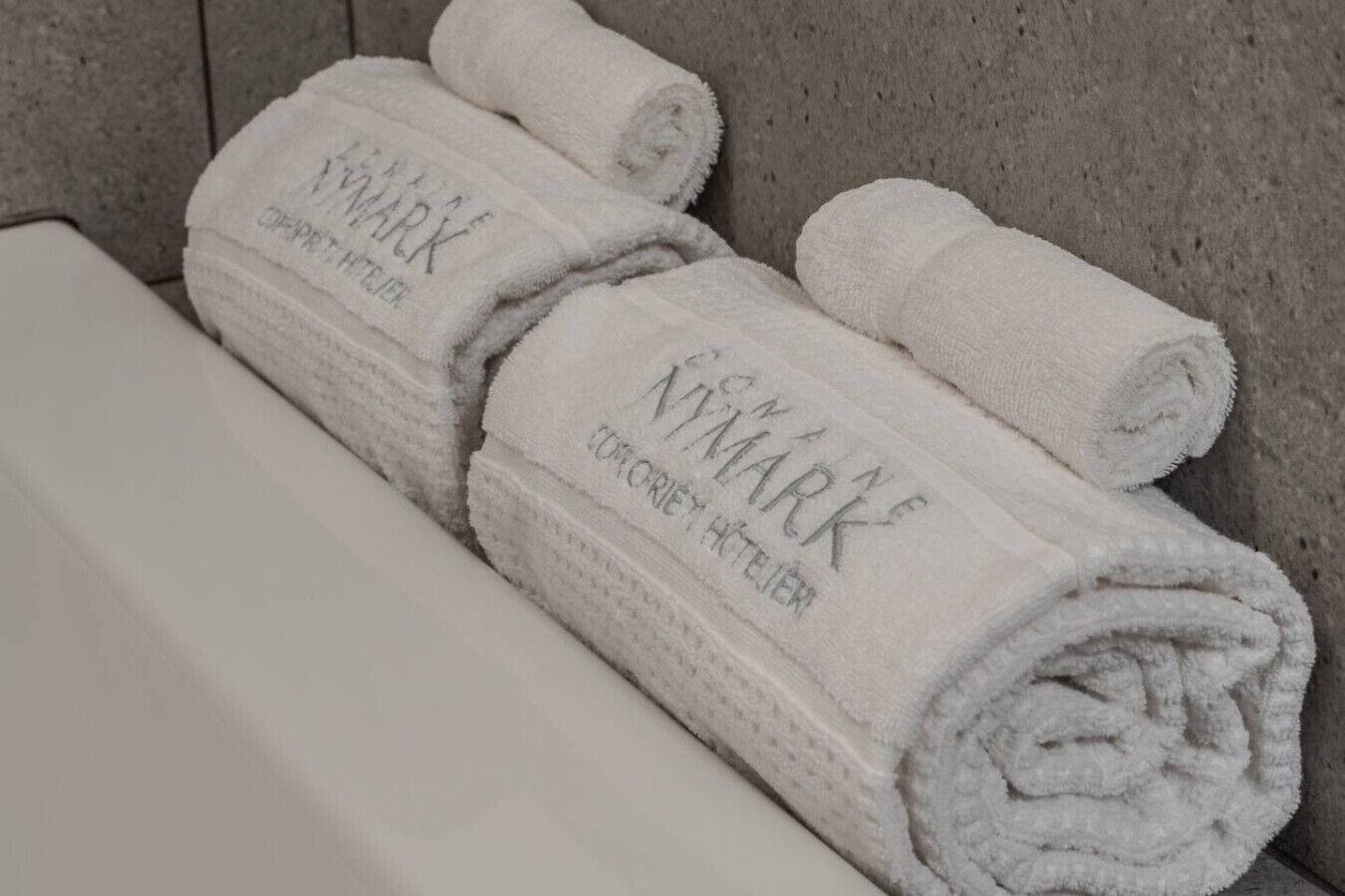 Drie opgerolde witte handdoeken met de tekst 'VIKIMARK' en 'HOTEL HETTER', op een witte ondergrond voor een grijze muur.