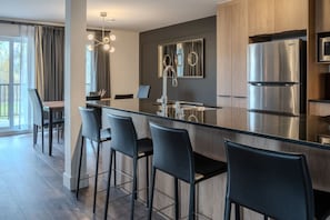 A modern kitchen with a central island, stainless steel appliances, and black bar stools. - Condo 2 chambres - Ski in Ski out St Sau #206 (Saint-Sauveur)