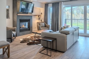 A modern living room with a fireplace, leather sofa, and a large window with curtains. - Condo 2 chambres - Ski in Ski out St Sau #206 (Saint-Sauveur)