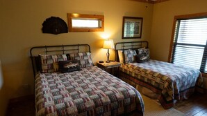 Una habitación con dos camas, cada una con colcha, una mesita de noche con lámpara, un cuadro enmarcado en la pared y una ventana con persianas.