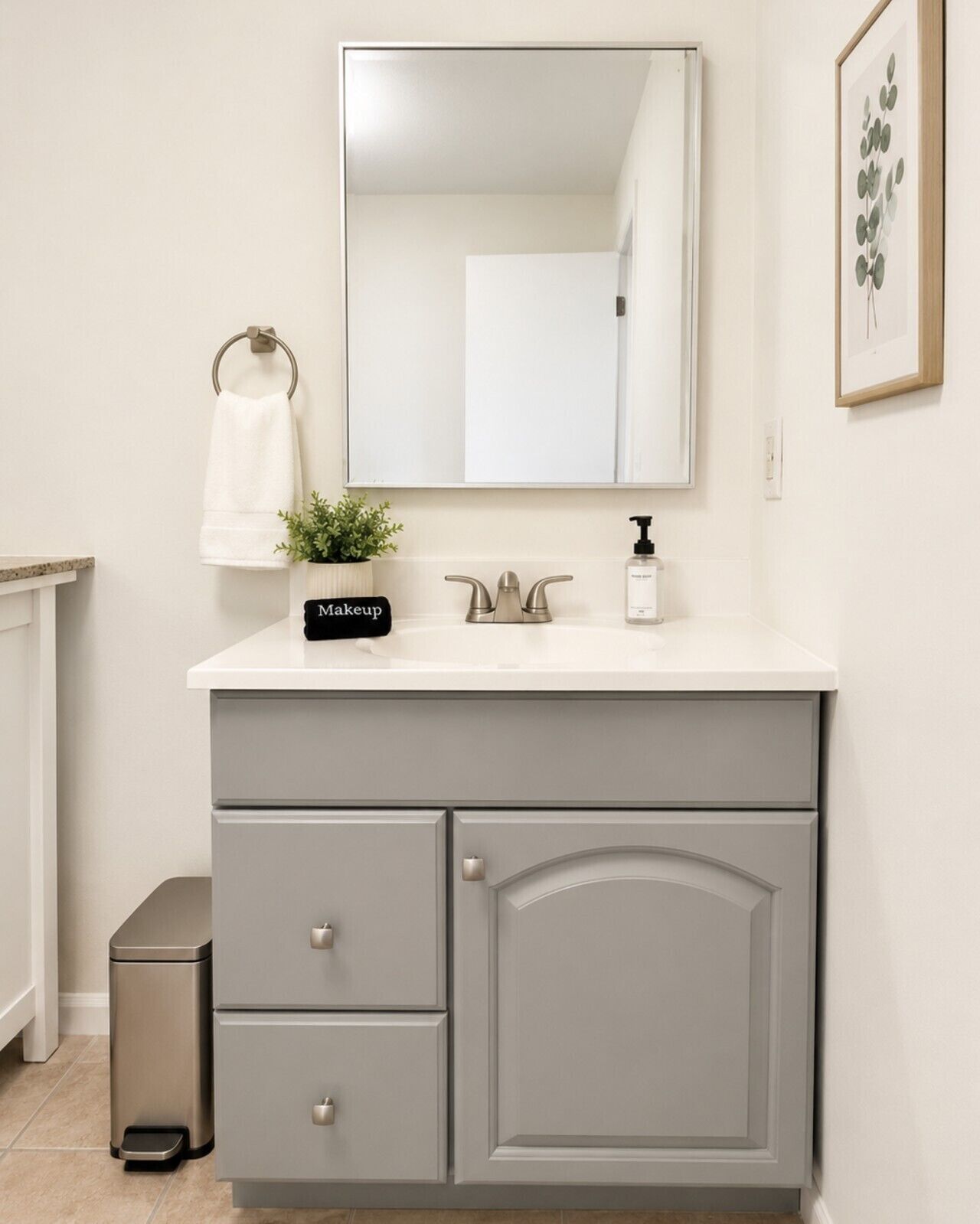 A bathroom with a white vanity, a mirror, a sink with a soap dispenser, a small potted plant, a towel ring, and a trash bin.