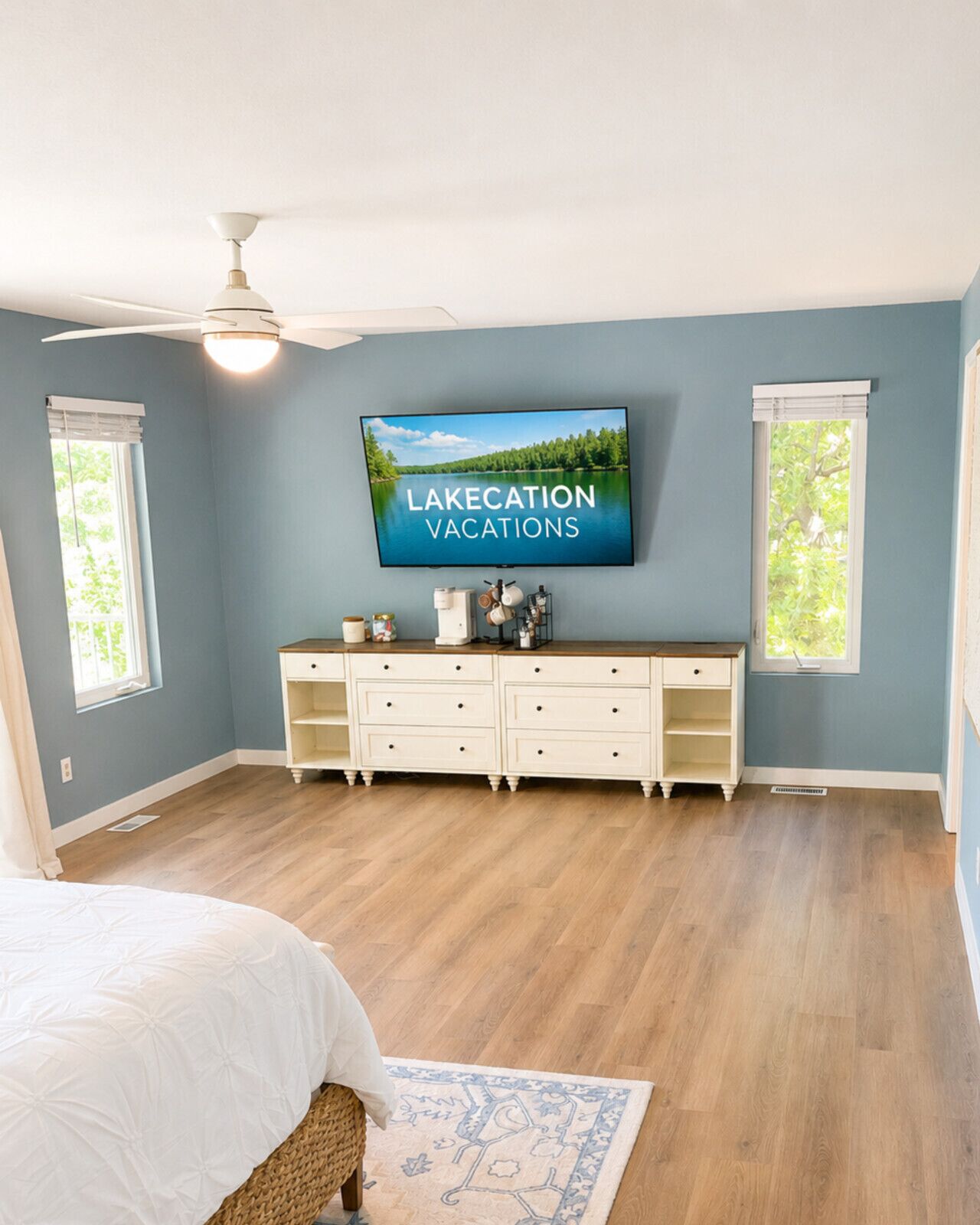 A bedroom with a bed, a dresser, a television displaying 'LAKECATION VACATIONS,' and a ceiling fan.