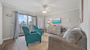 Una sala de estar con sillones azul verdosos, un sofá gris, un ventilador de techo, un televisor y vistas al exterior.