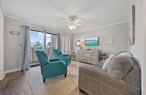 Ein Wohnzimmer mit türkisfarbenen Sesseln, einem grauen Sofa, einem Deckenventilator, einem Fernseher und Blick ins Freie.