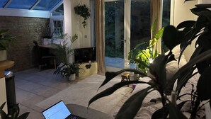 A modern living room with a large TV, a laptop, and a dining area with a table and chairs. There are several potted plants and a sliding glass door leading to a balcony. - Spacious Garden Flat- Walk to Wimbledon (London)