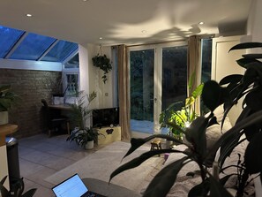 A modern living room with a large TV, a laptop, and a dining area with a table and chairs. There are several potted plants and a sliding glass door leading to a balcony. - Spacious Garden Flat- Walk to Wimbledon (London)