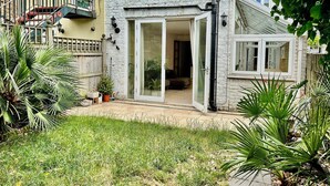 A residential building with a sliding glass door entrance, a small garden area with grass and potted plants, and a staircase leading to an upper level. - Spacious Garden Flat- Walk to Wimbledon (London)