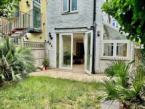 A residential building with a sliding glass door entrance, a small garden area with grass and potted plants, and a staircase leading to an upper level. - Spacious Garden Flat- Walk to Wimbledon (London)