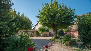 Une cour avec un chemin de gravier, entourée de bâtiments aux volets rouges, d’un grand arbre et de fleurs rouges.