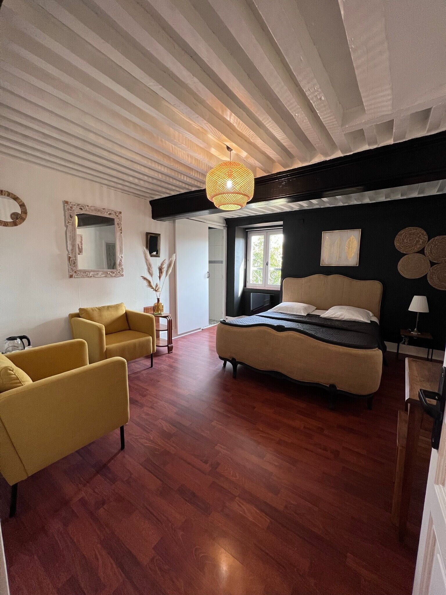 Une chambre à coucher avec un lit, deux fauteuils jaunes, un grand miroir et un luminaire suspendu.