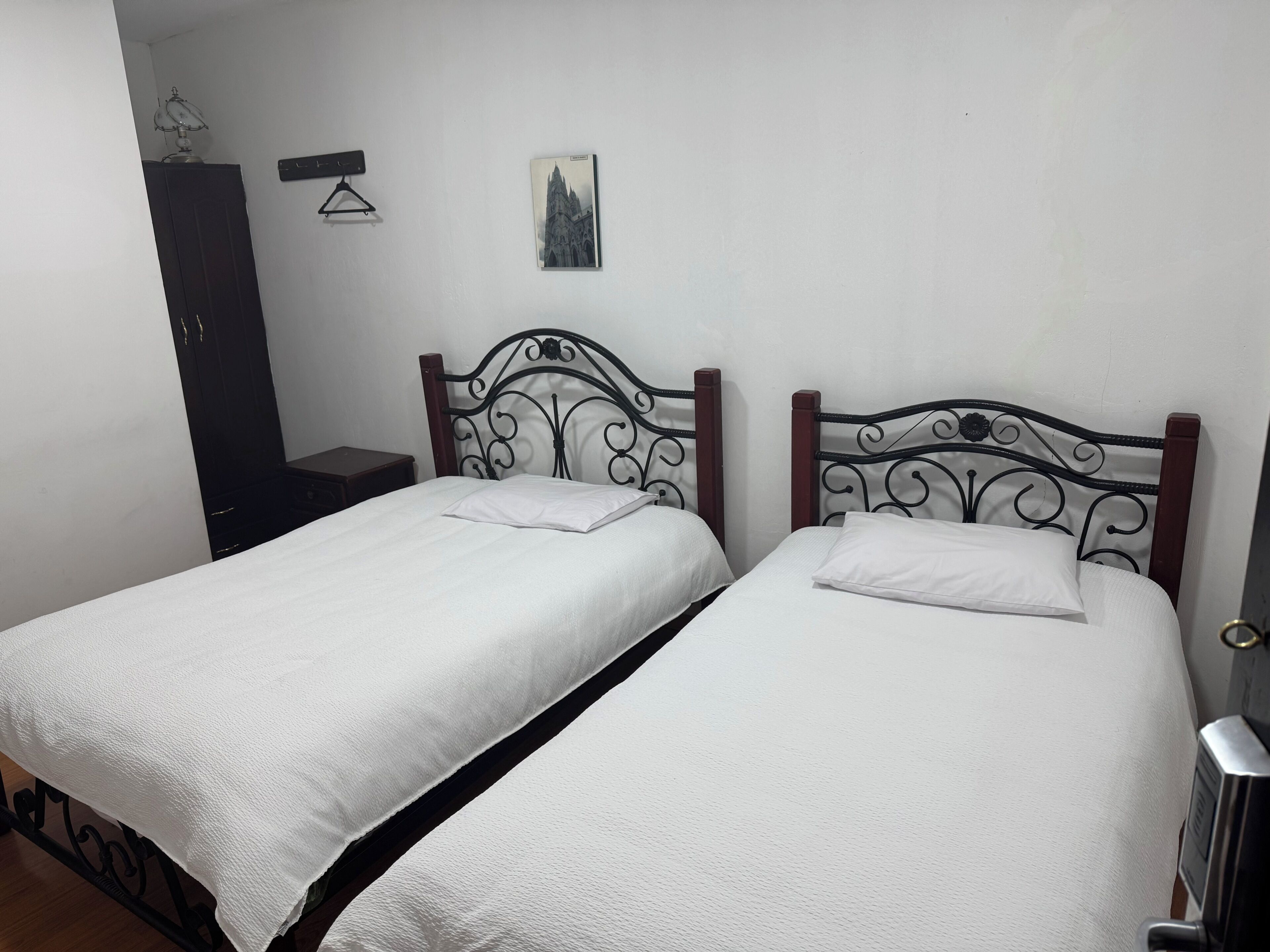 A room with two single beds, a wooden wardrobe, a small table, and a wall-mounted picture.