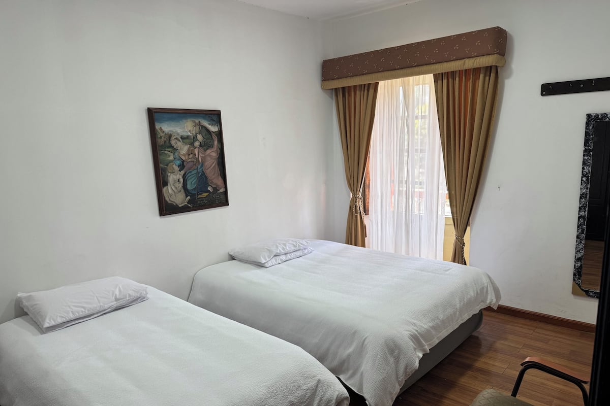 A room with two beds, a painting on the wall, a window with curtains, and a chair.