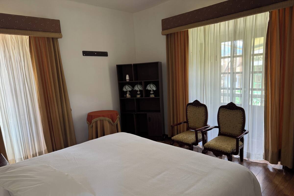 A hotel room with a large bed, two armchairs, a small table, a bookshelf, and a window with curtains.
