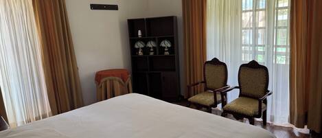 A hotel room with a large bed, two armchairs, a small table, a bookshelf, and a window with curtains.