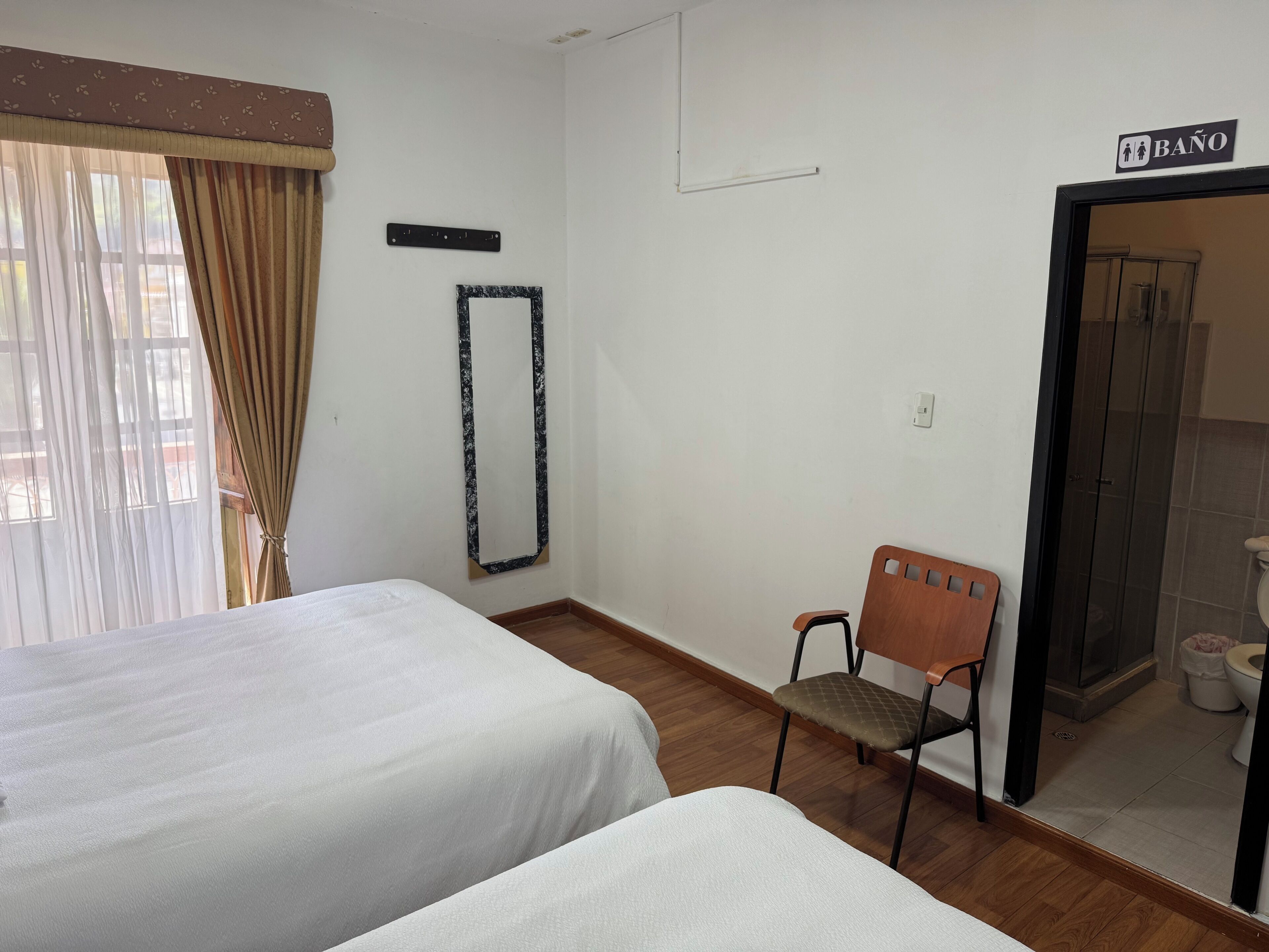 A hotel room with two beds, a chair, a mirror, a 'BAÑO' sign, and a bathroom visible through an open door.