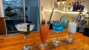 A bar area with three different drinks, a wooden counter, and shelves with bottles in the background.